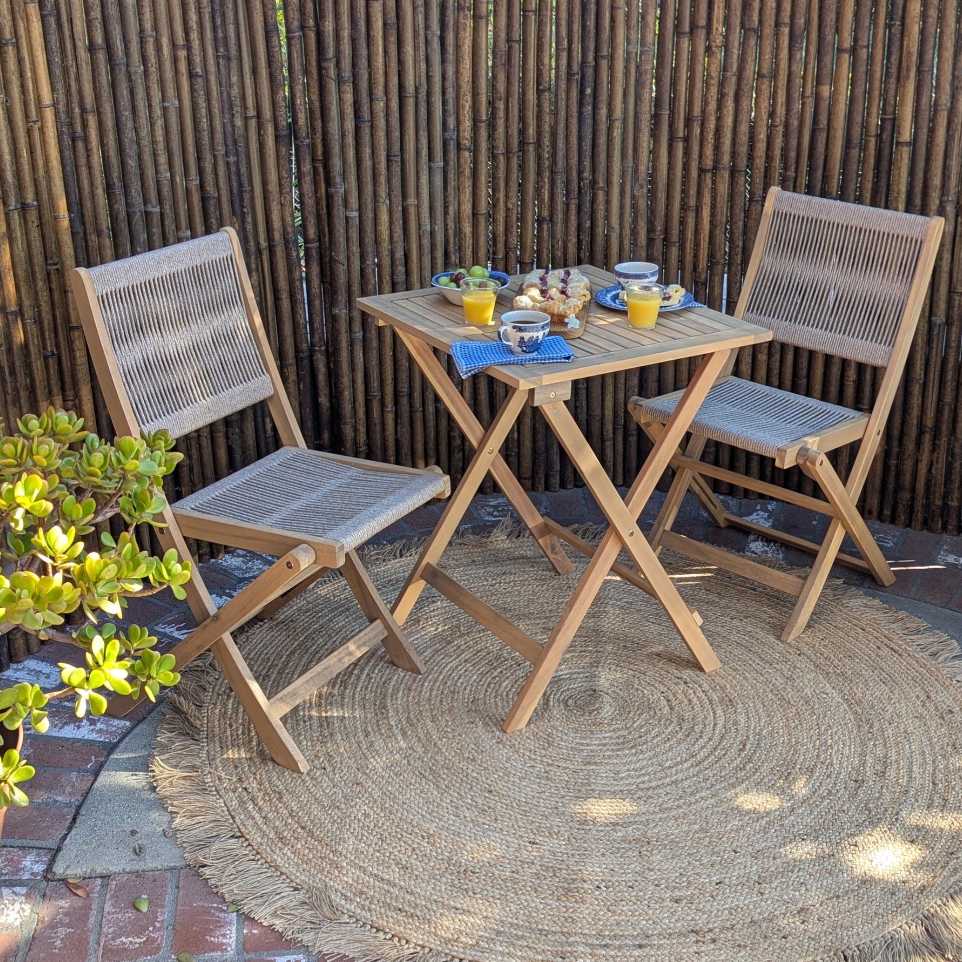 Outdoor patio set with wooden table and chairs on a woven rug against a bamboo fence.