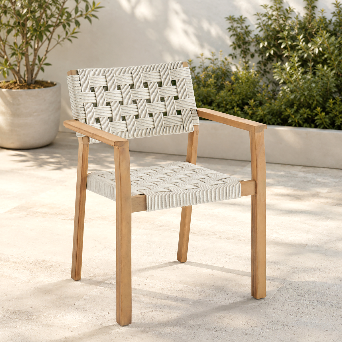 A modern outdoor armchair with a light natural wood frame and woven off-white rope seat and back, shown on a concrete patio with potted plants in the background.
