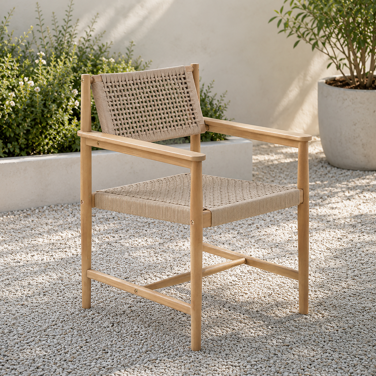 A light natural wood outdoor armchair with woven rope seats and backrest, shown on a gravel patio with potted plants in the background.