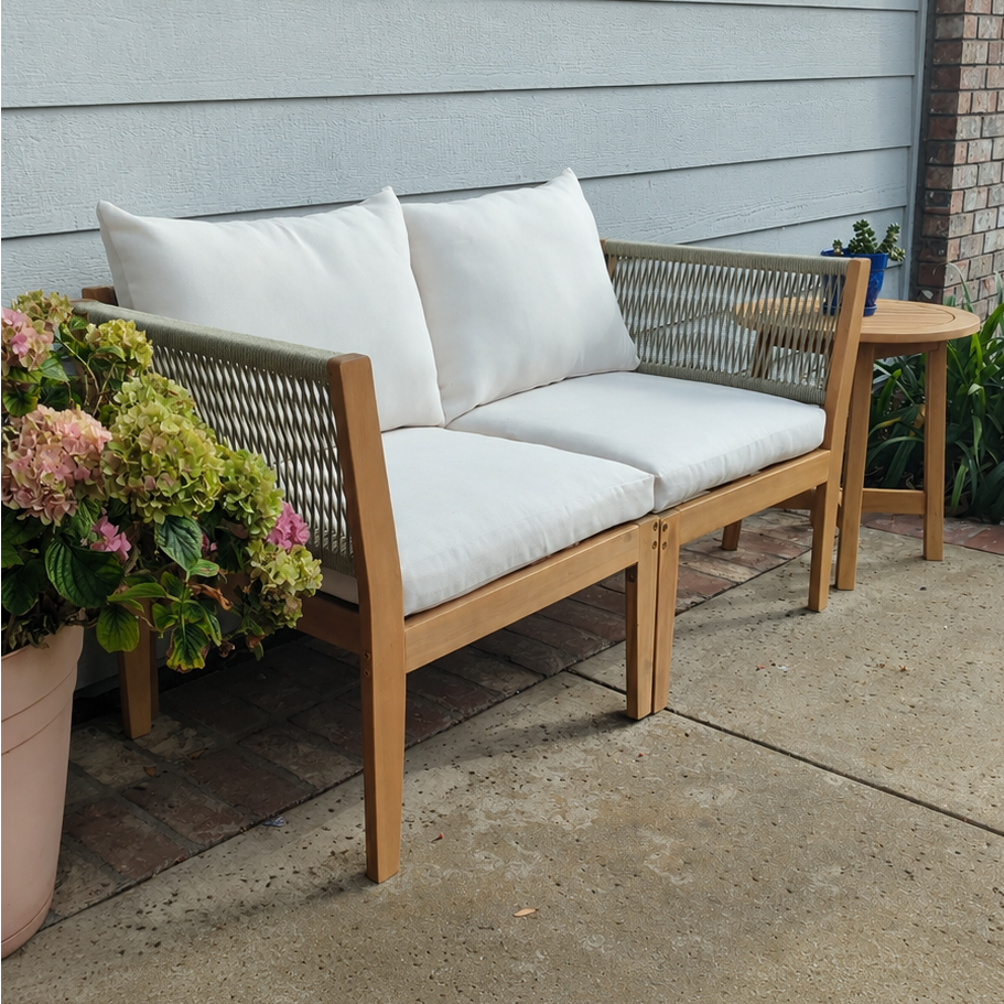 A wooden outdoor loveseat with woven backrest panels and white cushions, placed on a concrete patio in front of a house with light gray siding. A small matching side table and potted flowers are nearby.
