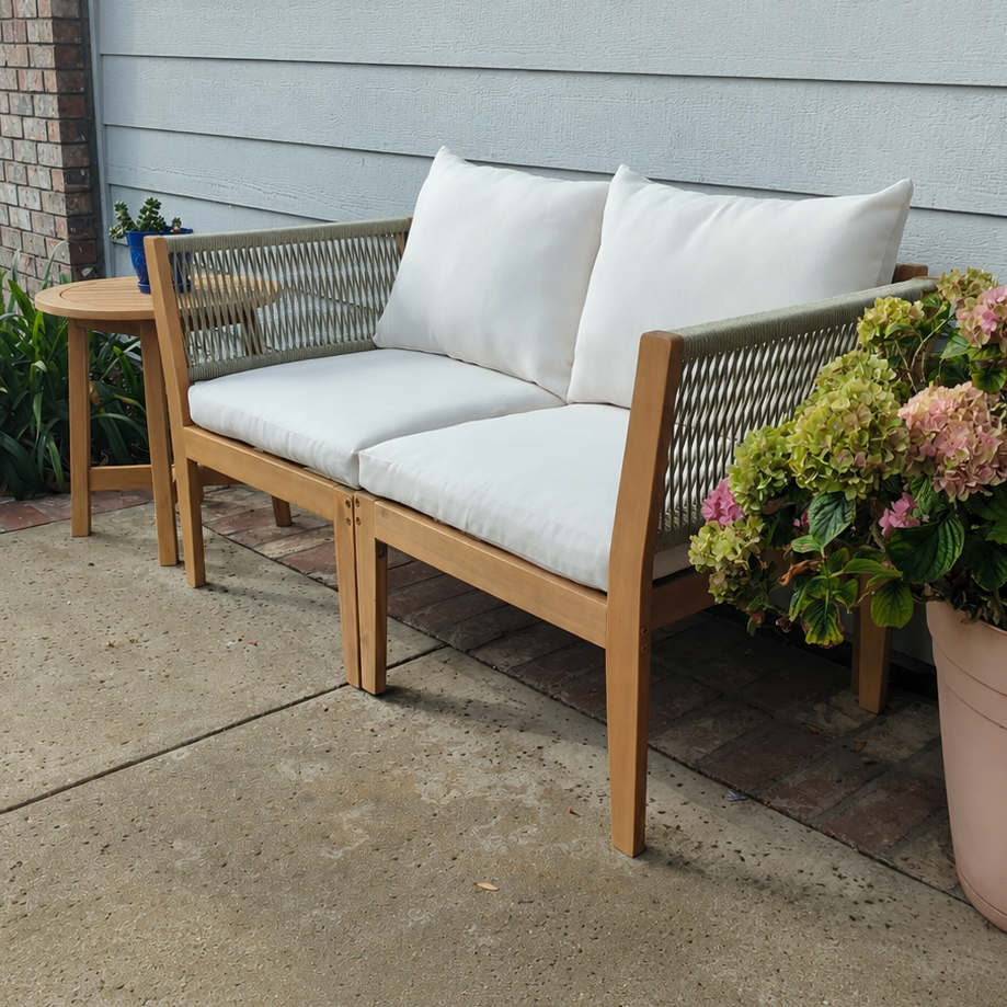 A wooden outdoor loveseat with woven backrest panels and white cushions, placed on a concrete patio in front of a house with light gray siding. A small matching side table and potted flowers are nearby.