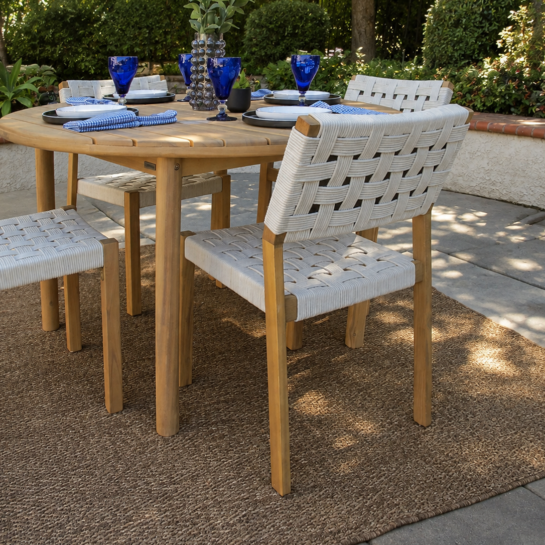 Outdoor dining set featuring a round natural wood table and four matching woven-back chairs, styled on a jute rug in a garden patio setting.